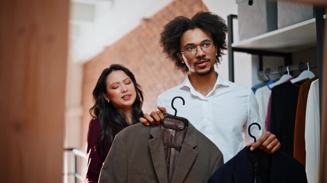 Multiethnic pair comparing blazer boutique. Man holding suit jacket smiling.