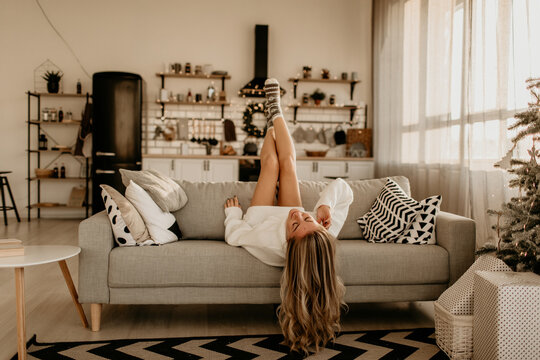 Young woman lying upside down on sofa near Christmas tree in New Year room, woman playfully raising legs near shelving, woman relaxing in cozy festive interior with grain, high quality photo