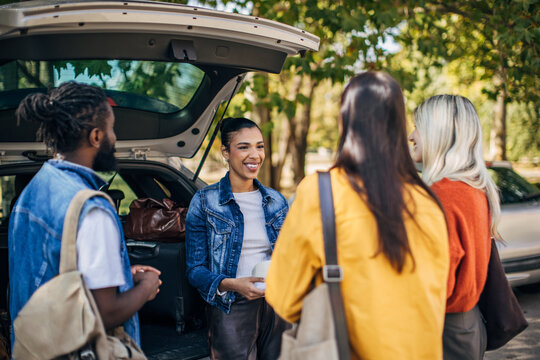 A group of diverse friends stands by an open car trunk, chatting and laughing as they prepare for a road trip. The warm sunlight filters through the trees, creating a joyful atmosphere for their adven