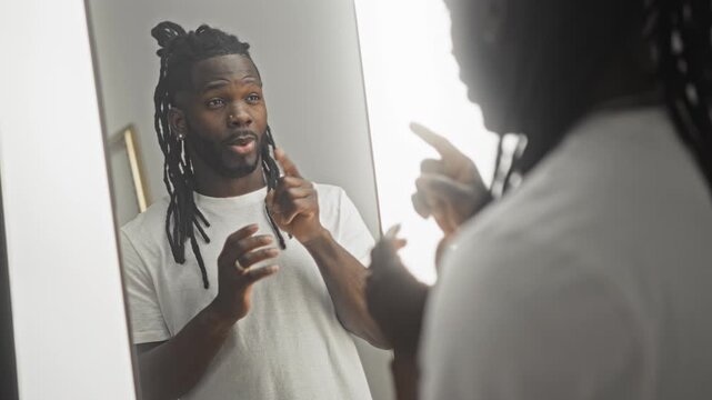 Young african american man in studio mirror forming ok sign with hands and speaking to his reflection while wearing white tshirt; introspection.
