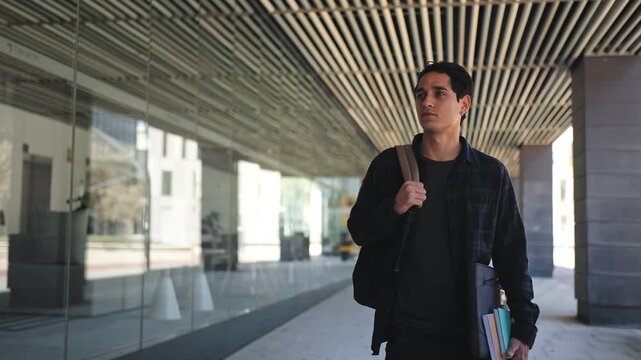 Young latin student carrying laptop books backpack walking outdoors near modern university building