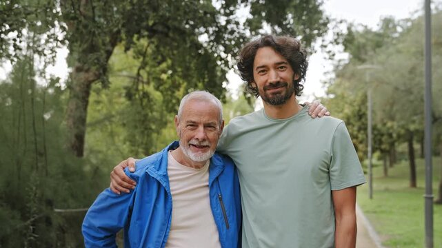 Portrait of senior father and adult son smiling at camera bonding with arms around shoulders wearing sportswear in park 