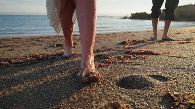 Low angle close up of barefoot woman in white lace dress walking on dark sand with red seaweed, sand falling from her feet. Man waits by the sea, rocky headland and waves behind. Warm evening light.