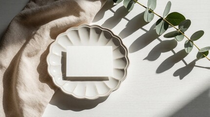Fototapeta premium Minimalist mockup features blank white card on scalloped ceramic plate. Eucalyptus branch, beige linen fabric rest on white wooden table. Natural sunlight creates soft shadows for wedding invitation