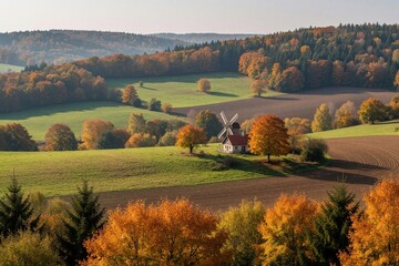 Obraz premium Old windmill stands in scenic autumn landscape. Colorful fall trees surround white building on rolling green hills. Rural farm field meets dense forest under bright sky. Beautiful seasonal view of