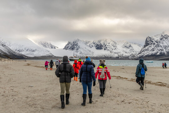 Expedition tourists walking across a remote Arctic beach toward a passenger ship with glacier and snow-covered mountains ahead, Spitsbergen, Svalbard, Norway