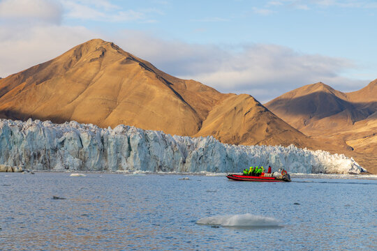 Arctic expedition boat carrying travelers across cold fjord water beneath a massive blue glacier and golden mountain slopes, Spitsbergen, Svalbard, Norway