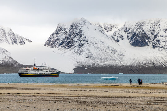 Expedition ship and tourists on a remote Arctic beach with floating iceberg and dramatic snow-covered mountains in summer, Spitsbergen, Svalbard, Norway