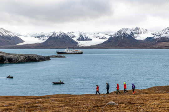 Group of tourists trekking along remote tundra shoreline while zodiac boats cross the fjord beneath snow-covered mountain ridges, Ny-London, Blomstrandhalvoya, Spitsbergen, Svalbard, Norway