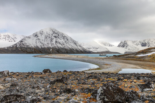 Wide Arctic summer landscape with rocky tundra foreground, calm fjord water, distant hikers, ship and snow-covered mountains, Spitsbergen, Svalbard, Norwa