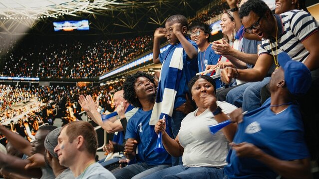 Young child fan stands among cheering blue supporters during a soccer match in a packed stadium, sharing authentic family fun as a Black family cheers together and supports the blue team.