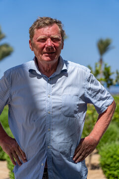 Mature man in blue striped short sleeve shirt standing outdoors with hands on his hips against bright sunny background with palm trees and green foliage.