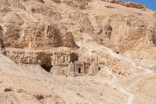 Ancient Egyptian tomb entrance carved into rocky desert hillside under clear blue sky with path leading to structure.