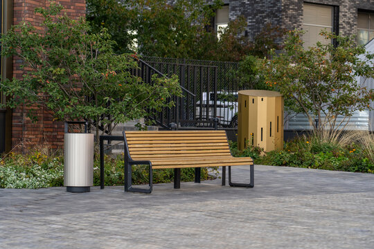 Wooden park bench sits on paved courtyard next to green bushes, metal trash can, and modern building facade in urban outdoor space during daytime.
