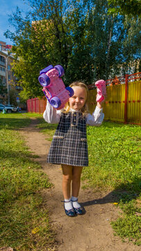Young girl holding two pink toy vehicles, car and remote control, while standing outdoors on sunny day.