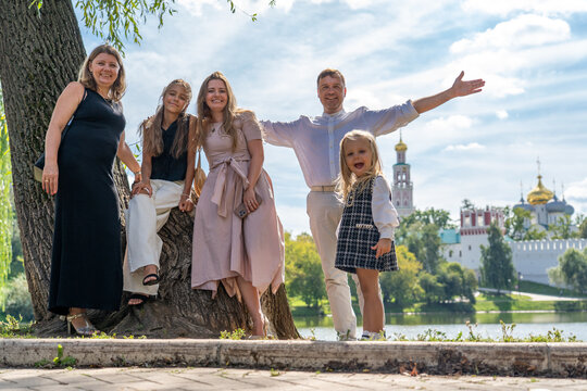 Happy multi-generation family stands together outdoors near large tree by lake, with cathedral and park in background on sunny day.