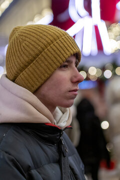 Profile of young man wearing mustard yellow beanie and puffer jacket against background of blurred festive lights.