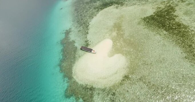 FPV aerial view of a small wooden boat anchored on a white sandbar surrounded by turquoise coral reefs and deep blue sea near Labuan Bajo in Komodo, East Nusa Tenggara, Indonesia.