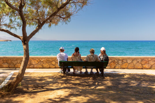 People sitting on a bench under a tree and looking at the turquoise Mediterranean Sea along the coast of Chania, Crete, Greece.