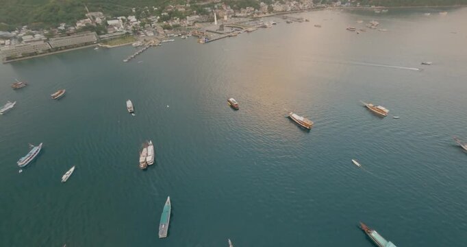 FPV aerial view of Labuan Bajo harbor with numerous boats anchored in the clear turquoise waters of the coast in Komodo, East Nusa Tenggara, Indonesia.