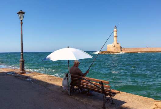 Local fisherman sitting under an umbrella by the sea with the historic Venetian lighthouse in Chania, Crete, Greece.