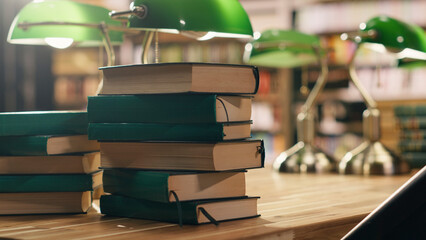 Stacked books sit on a desk in a quiet university library with warm lighting and a green lamp, surrounded by bookshelves and study materials that create an empty academic reading and study space. © DC Studio