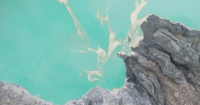 FPV aerial view of Ijen Volcano crater lake with turquoise water and rugged volcanic rock formations in East Java, Indonesia.