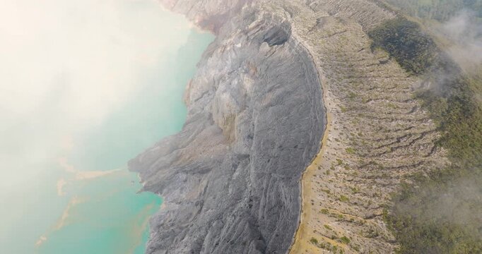 FPV aerial view of Ijen Volcano crater lake with turquoise water and rocky crater rim surrounded by mist, East Java, Indonesia.