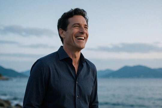 Middle aged caucasian man laughs outdoors near sea. Happy smiling male in navy striped shirt enjoys evening breeze on hilltop. Mature person feels joy looking away towards mountains, ocean view