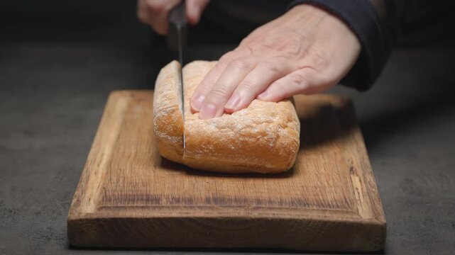 chef slices ciabatta bread loaf on wooden cutting board on black tabletop. Preparation of Italian tomato and ricotta sandwich; food closeup, healthy eating