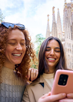 Vertical. Young only women friend using mobile phone in hands in front of Sagrada Familia in Barcelona. Beautiful caucasian girls at European city enjoying vacation happy smiling. Fun tourism people