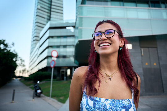 Portrait of a cheerful young woman with red hair and glasses, smiling brightly in a modern city environment, showcasing confidence and urban lifestyle