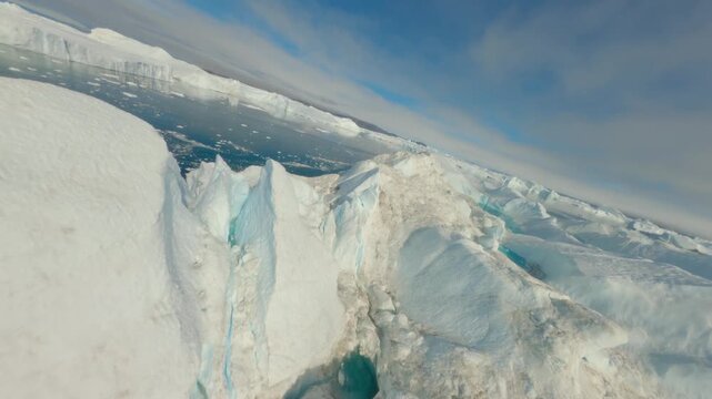 FPV aerial view of a massive iceberg floating in the deep blue waters of the Ilulissat Icefjord, surrounded by smaller ice fragments under a clear sky in Ilulissat, Avannaata, Greenland.