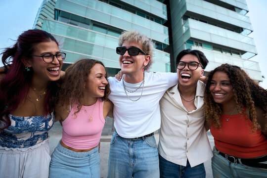 Diverse group of young friends standing with their arms around each other and laughing outdoors. Candid urban scene conveying friendship, inclusion, unity and positive youthful energy.