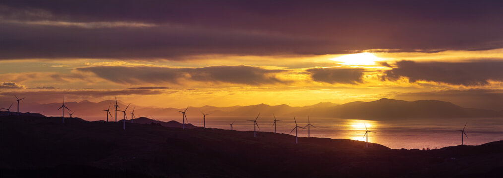 Wind turbines silhouetted on hills at sunset over Cook Strait, Mill Creek wind farm, Wellington, New Zealand, panorama. South Island Te Waipounamu in distance.