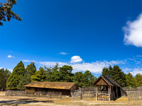 Rural farmyard with wooden buildings and fences. Green evergreen trees surround the open area under a blue sky with clouds.