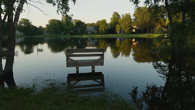 Suburban pond overflowing onto a wooden bench and surrounding grassy shoreline after heavy rainfall, showing localized flooding and high water levels near residential homes.