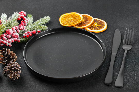 Christmas table setting with empty black plate, black cutlery, spruce branch, dried orange slices, glass and wrapped gifts on dark background. Template, mockup with festive holiday atmosphere.
