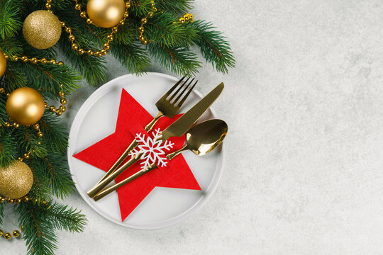 Holiday table setting with cutlery arranged in red star shaped holder decorated with snowflake on white plate, Christmas tree branch with godlen balls on light gray textured background, copy space