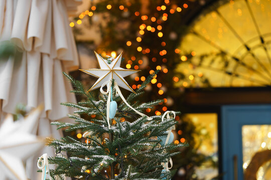 Christmas tree decorated with white star, ornaments and warm fairy lights, displayed outdoors with festive illuminated, light bokeh background and snowy branches. Holiday atmosphere, aesthetic mood