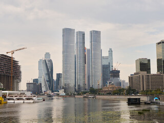 Fototapeta premium Modern city skyline with glass skyscrapers along riverfront, construction cranes at building site, boats docked near promenade, and overcast clouds