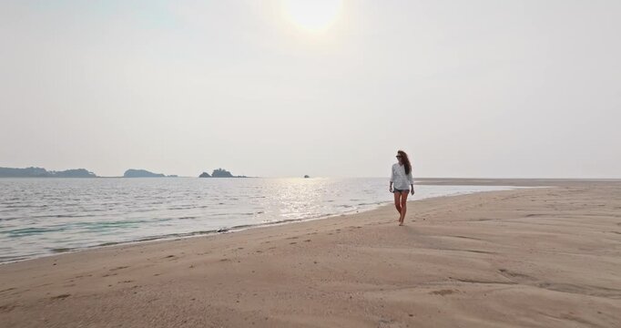 Young tourist woman walking barefoot on a remote tropical beach, enjoying peaceful solitude during a sunny vacation in Ko Lanta, Thailand, with clear sky and sea reflecting the sun. Travel, relax