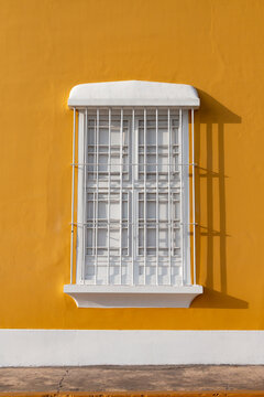 Colonial window with bright yellow wall with white window and door. Window features metal bars and a simple ledge. Door is closed, slightly ajar, with clean lines. Sunlight casts sharp shadows across 