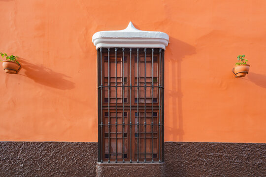Warm orange wall with dark metal window. White trim tops the window's frame. Potted plants flank the window, adding life. Solemn stillness, captured in quiet architectural detail. Pueblo Libre, Lima, 
