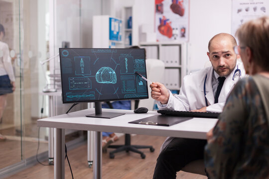 Doctor points at a digital brain imaging monitor while explaining results to an older woman patient during a face-to-face medical consultation.