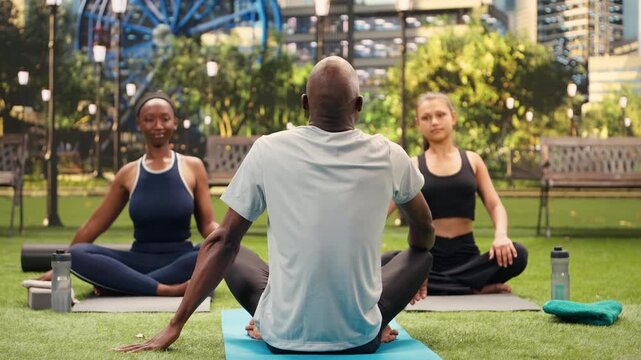 Yoga instructor faces participants seated cross-legged on mats in city park, guiding outdoor meditation and mindful deep breathing while women continue a calm Pilates wellness practice together.