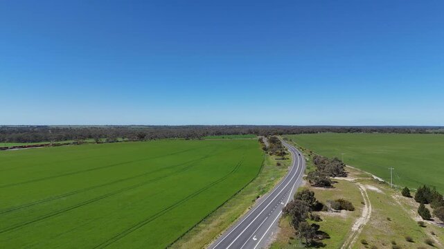 Rising aerial drone shot over Western Highway near Dimboola, Victoria, Australia, revealing a long freight train moving along the railway line parallel to the highway across expansive green cropland&nbsp;