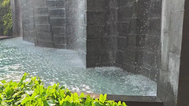 Modern artificial waterfall with stone wall and green plants in clear pool water