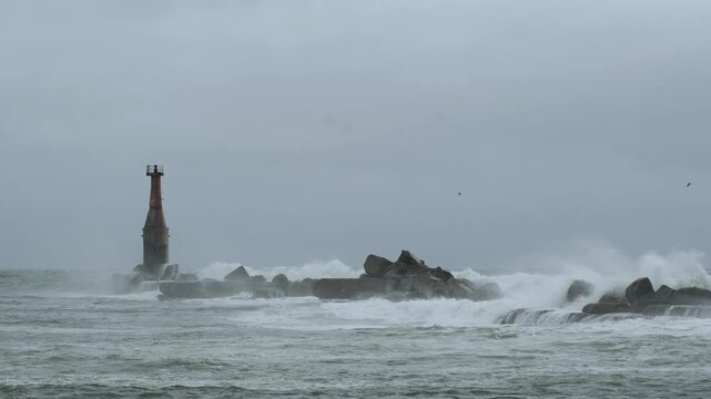 Raging sea waves crash against the old lighthouse and the stone breakwater.