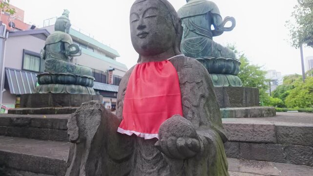 A small stone Jizo statue with a red bib sits before larger bronze Buddhas at Zojoji Temple in Minato, Tokyo, Japan, representing devotion.